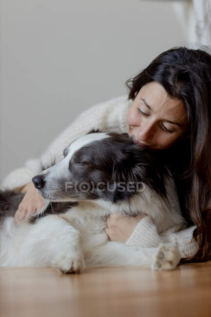 Fürsorgliche Hündin im Wollpullover umarmt lustigen Border Collie Hund, während sie gemeinsam auf dem Holzboden liegt — Stockfoto