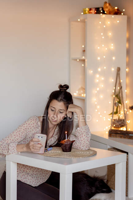 Нещаслива жінка у повсякденному одязі сидить за столом з смачним кексом і читає повідомлення на мобільному телефоні під час святкування дня народження лише під час карантину. — стокове фото