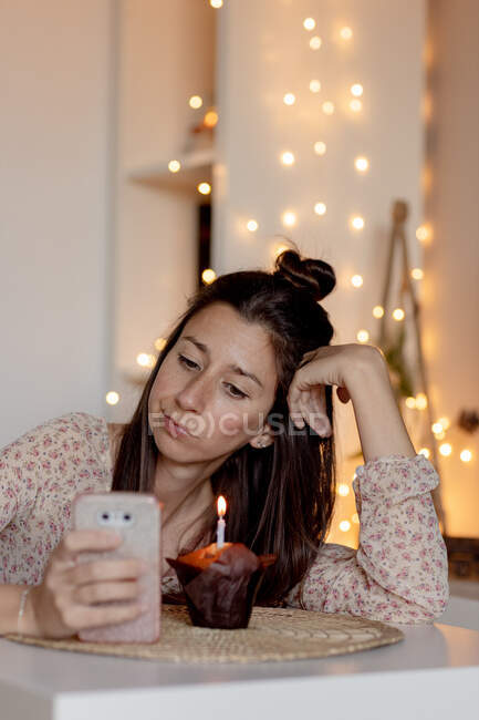 Femme malheureuse en tenue décontractée assise à table avec un délicieux muffin et lisant des messages sur son téléphone portable tout en célébrant son anniversaire seul pendant la quarantaine — Photo de stock