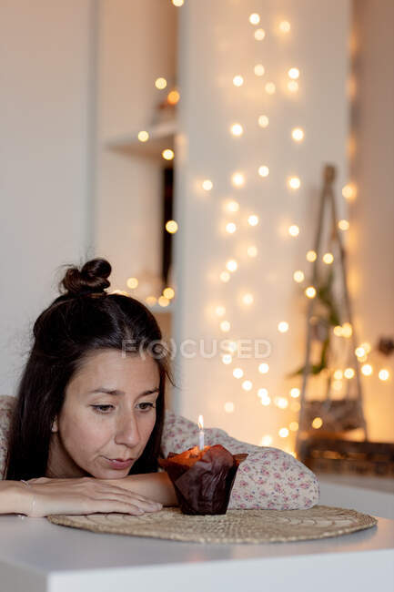 Сумна жінка в повсякденному одязі сидить за столом з мафіном і святкує день народження на самоті, перебуваючи вдома під час карантину — стокове фото