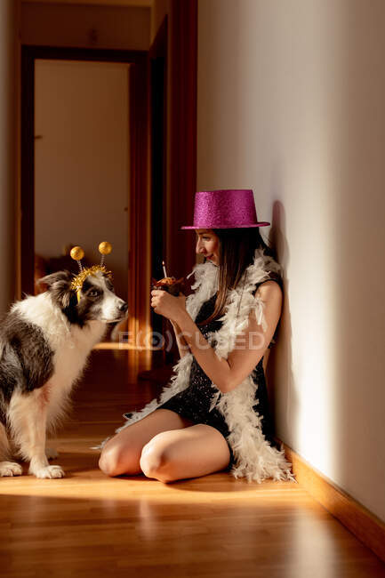Vista lateral de la mujer sonriente con sombrero de fiesta y vestido sentado en el suelo con magdalena y celebrando el cumpleaños junto con el perro durante la pandemia covid 19 - foto de stock