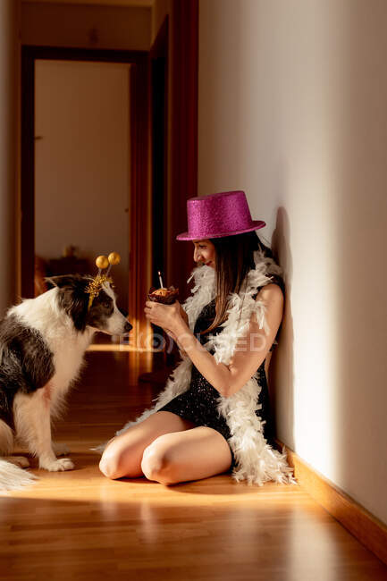 Vista laterale di una donna sorridente che indossa cappello e vestito da festa seduta sul pavimento con muffin e festeggia il compleanno insieme al cane durante la pandemia covid 19 — Foto stock