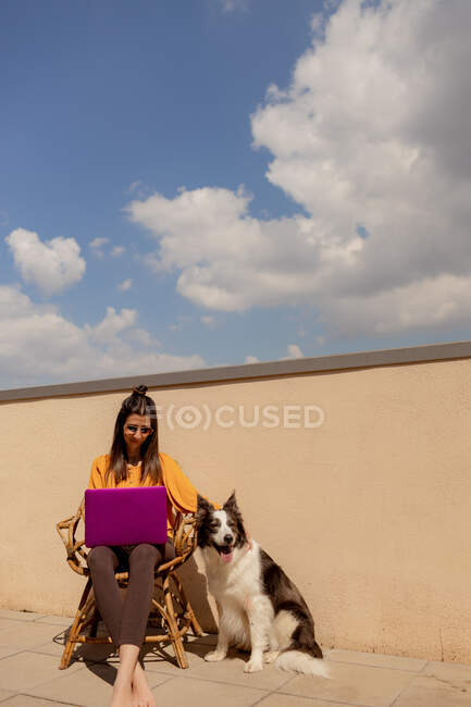 Brünette Frau in lässigem Outfit sitzt auf Stuhl und blättert im lila Netbook, während sie arbeitet und während der Quarantäne die Sonne auf der Terrasse des Hauses genießt — Stockfoto