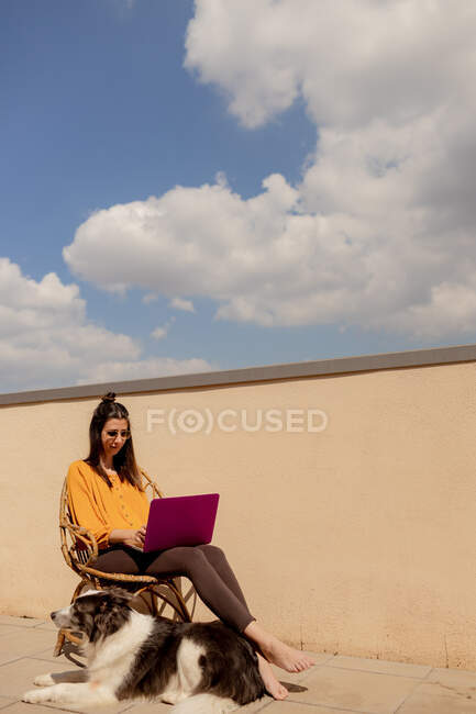 Bruna donna in abito casual seduto sulla sedia e la navigazione netbook viola mentre si lavora e godersi il sole in terrazza di casa durante la quarantena — Foto stock