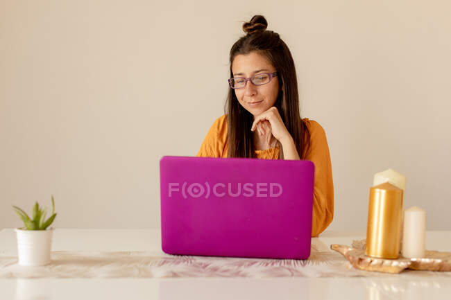 Mujer joven en traje casual y anteojos sonriendo y mirando a la computadora portátil mientras se sienta en la mesa en la habitación acogedora - foto de stock