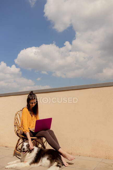 Brunette woman in casual outfit sitting on chair and browsing purple netbook while working and enjoying sunshine in terrace of house during quarantine — Stock Photo