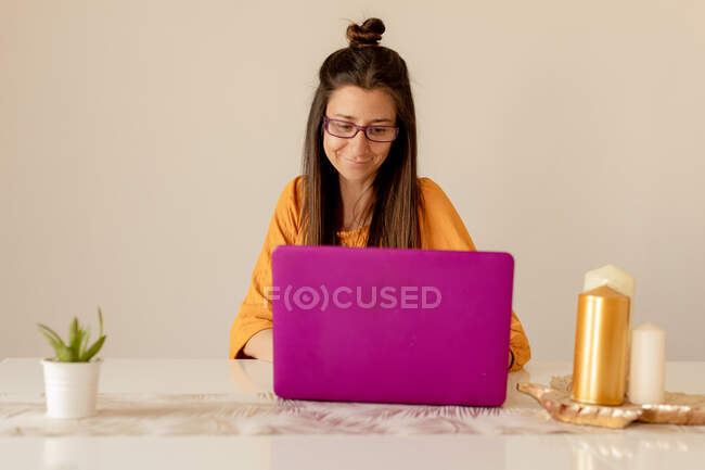 Молода жінка в повсякденному вбранні і окулярах посміхається і дивиться на ноутбук, сидячи за столом у затишній кімнаті — стокове фото