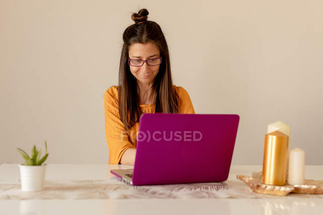 Giovane donna in abito casual e occhiali sorridente e guardando il computer portatile mentre seduto a tavola in camera accogliente — Foto stock