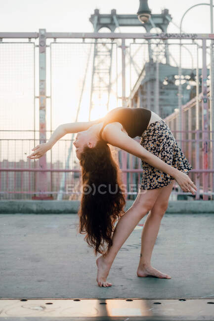 Вид сбоку на молодую грациозную танцовщицу в повседневной юбке, которая, стоя босиком на мосту, освещена сзади — стоковое фото