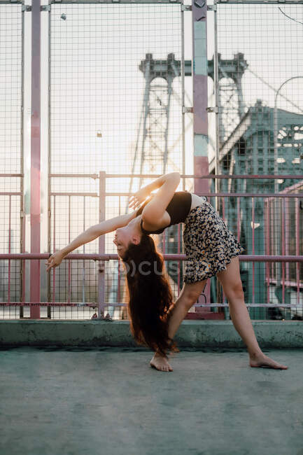 Side view of young graceful female dancer in casual skirt performing back bend while standing barefoot on bridge in back lit — Stock Photo