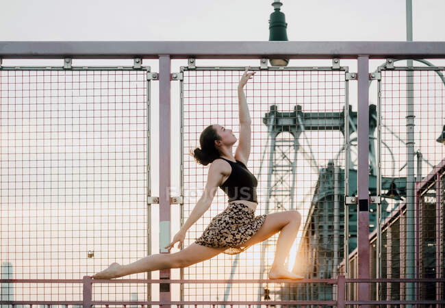 Vista lateral de ángulo bajo de una mujer serena que usa ropa de verano haciendo yoga en pose de zancada creciente mientras se equilibra en la barandilla de metal y mira hacia arriba - foto de stock