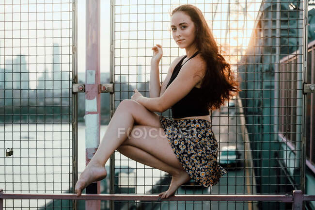 Вид збоку витонченої жінки, що балансує на металевому паркані міського мосту, сидячи босоніж на тлі міського пейзажу і дивлячись — стокове фото