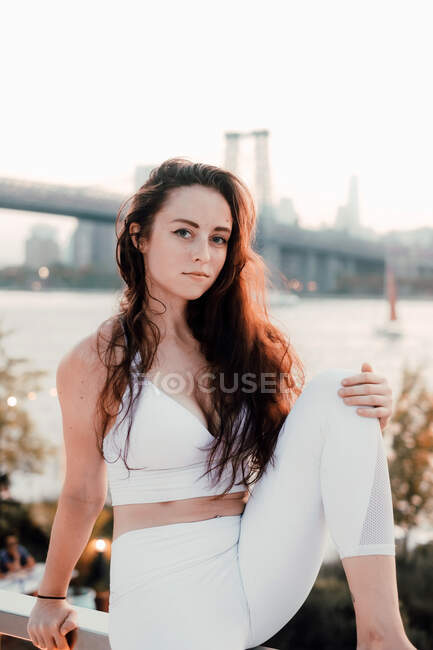 Великолепная женщина с длинными волосами в яркой одежде сидит на фоне моста и реки, расслабляясь и глядя в сторону — стоковое фото