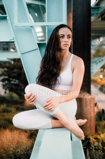 Мрія жінка у спортивному вбранні сидить на металевому балці міського мосту, насолоджуючись міським пейзажем і озираючись — стокове фото
