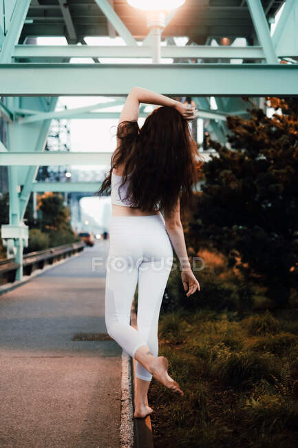 Jeune femme mince en haut de sport et leggings debout pieds nus sur le trottoir et levant le bras gracieusement dans la ville du soir — Photo de stock