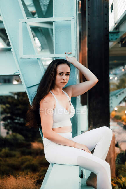 Gorgeous woman with long hair wearing bright clothes sitting on background of bridge and river while relaxing and looking away — Stock Photo
