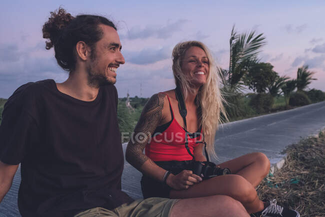 Содержит пару путешествующих хипстеров, сидящих на обочине дороги вечером и наблюдающих закат, расслабляясь и отводя взгляд — стоковое фото