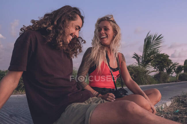 Zufriedenes Paar reisender Hipster, die abends am Straßenrand sitzen und den Sonnenuntergang beobachten, während sie sich entspannen und wegschauen — Stockfoto