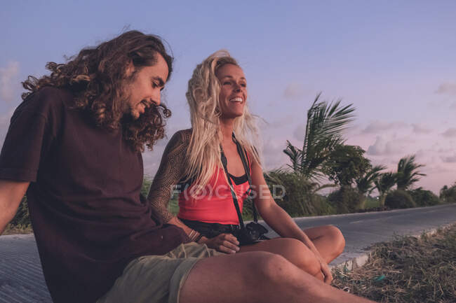 Contenu couple de hipsters itinérants assis sur le bord de la route le soir et regarder le coucher du soleil tout en se relaxant et en regardant loin — Photo de stock