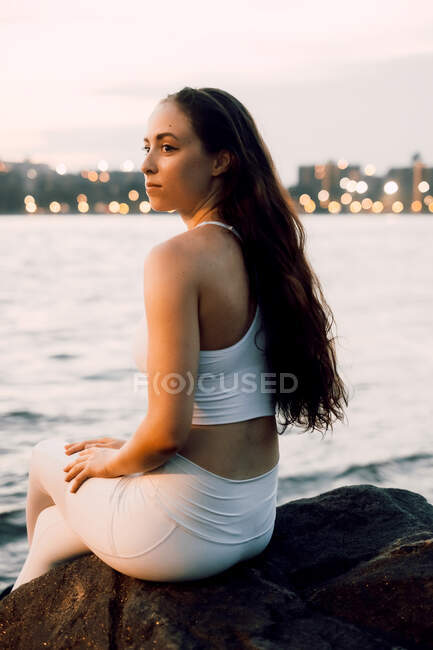 Vista lateral da mulher concentrada em activewear sentado em pedra à beira-mar enquanto pratica ioga no fundo da paisagem urbana da noite — Fotografia de Stock