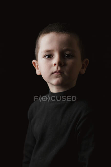 Portrait d'un petit garçon réfléchi regardant la caméra pendant la prise de vue en studio sur fond noir — Photo de stock