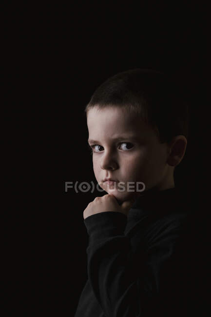 Retrato del niñito reflexivo mirando a la cámara durante la toma de fotos de estudio contra el fondo negro - foto de stock