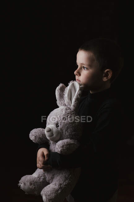 Студійний знімок спокійного маленького хлопчика в повсякденному светрі обіймає улюбленого плюшевого сірого кролика, дивлячись в очі і позуючи на чорному тлі — стокове фото