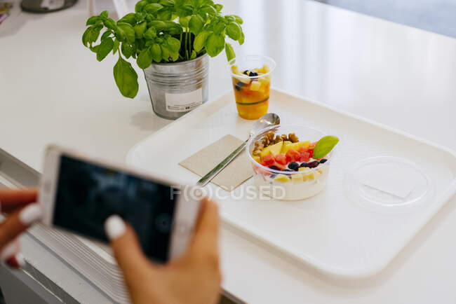 Desde arriba de mujer irreconocible tomando fotos con teléfono inteligente mientras está sentado en la mesa en el restaurante y comiendo deliciosos desiertos de frutas saludables en el plato blanco - foto de stock