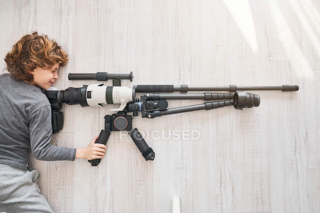 Зверху хлопчик грає з аксесуарами для професійної фотокамери, розташованої на підлозі у формі снайперської гвинтівки — стокове фото