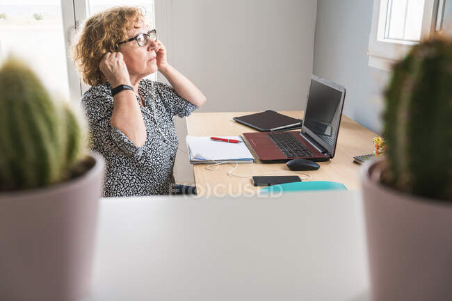 Вид сбоку на взрослую женщину в повседневной одежде, работающую на ноутбуке в наушниках в комнате, украшенной кактусами в керамических горшках — стоковое фото