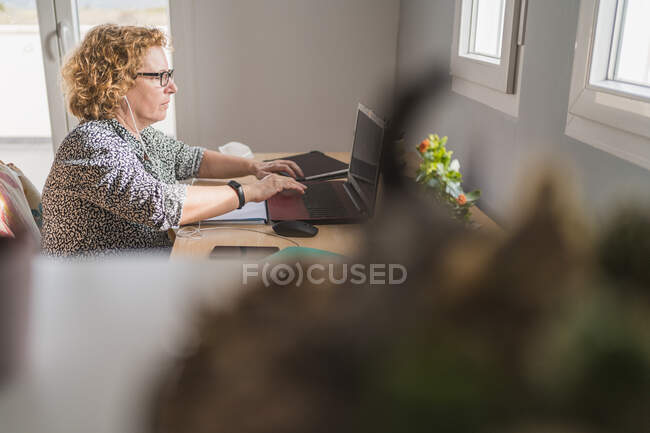 Vista lateral de la mujer adulta en ropa casual que trabaja en el ordenador portátil en auriculares en la habitación decorada con cactus en macetas de cerámica - foto de stock