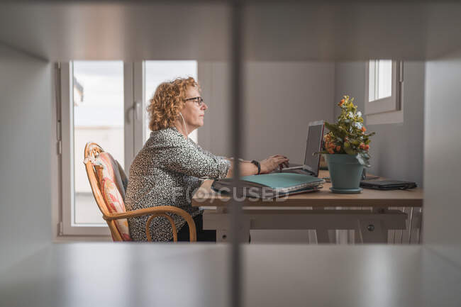 Seitenansicht einer erwachsenen Frau in Freizeitkleidung, die in einem Raum mit Kakteen in Keramiktöpfen auf einem Laptop in Kopfhörern arbeitet — Stockfoto