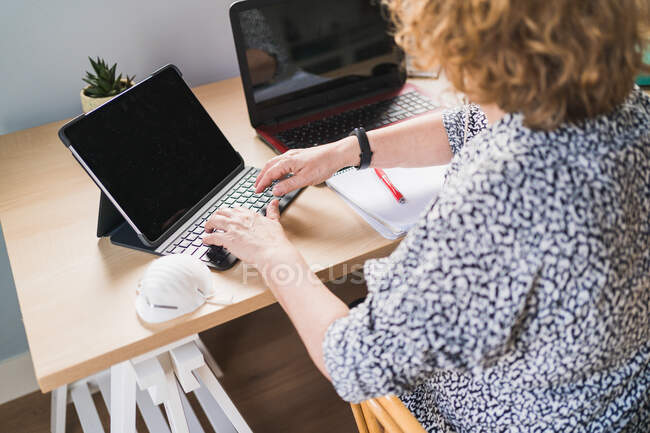 From above view of anonymous female freelancer in casual clothes typing on keyboard while working with laptops at wooden table at home — Stock Photo