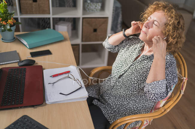 Side view of focused adult woman in casual clothes enjoying music in earphones with closed eyes while resting during work on laptop and notebook — Stock Photo