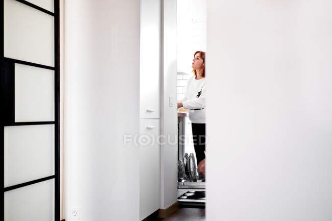 Vista lateral da dona de casa pensativa em roupas casuais carregando máquina de lavar louça com utensílios de mesa enquanto passa o fim de semana em casa — Fotografia de Stock