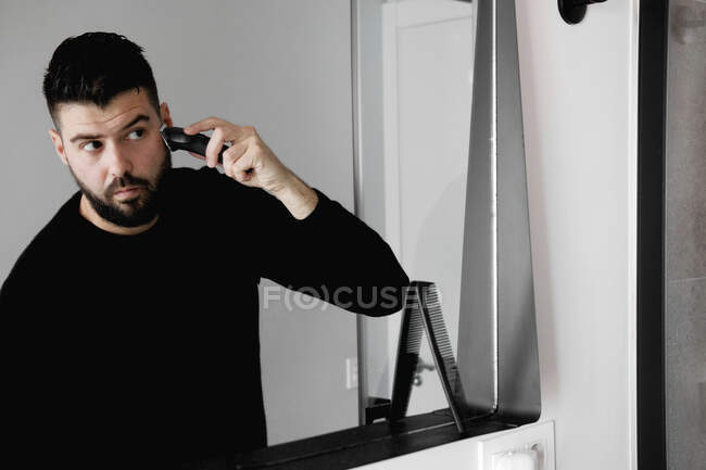 Серйозний красивий чоловік використовує електричну бритву для догляду, стоячи в сучасній ванній кімнаті і дивлячись у дзеркало — стокове фото
