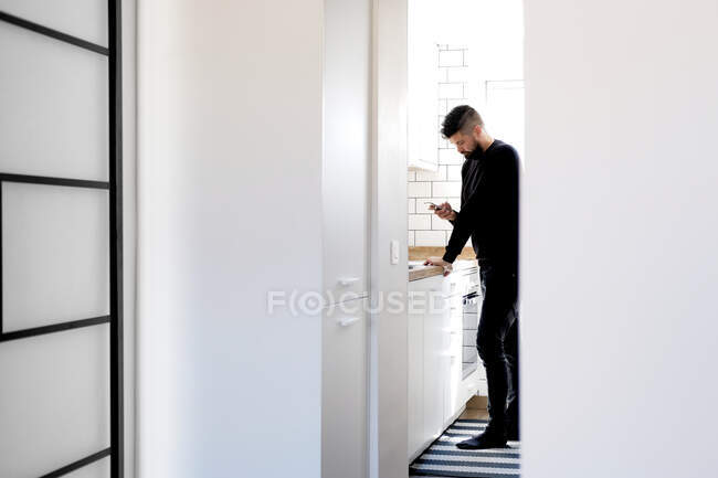 Side view of stylish man in casual outfit standing and chatting online on social media while leaning on countertop in bright kitchen interior — Stock Photo