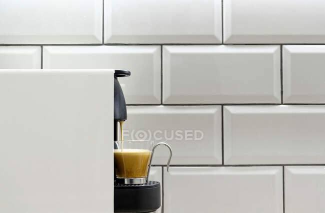Modern pod coffee maker pouring hot espresso into glass cup on background of ceramic tile on kitchen wall — Stock Photo