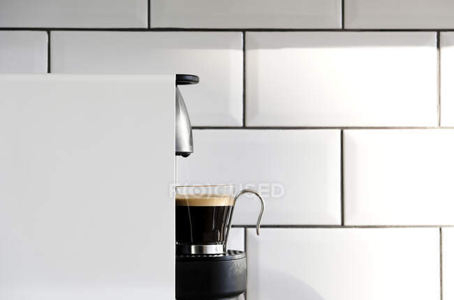 Современный стручок кофеварка наливая горячий эспрессо в стеклянную чашку на фоне керамической плитки на стене кухни — стоковое фото