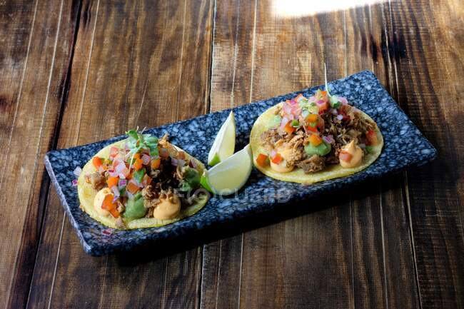High angle of delicious taco made of corn tortilla and meat garnished with chopped fresh vegetables and sauces while served on plate with lime wedges — Stock Photo