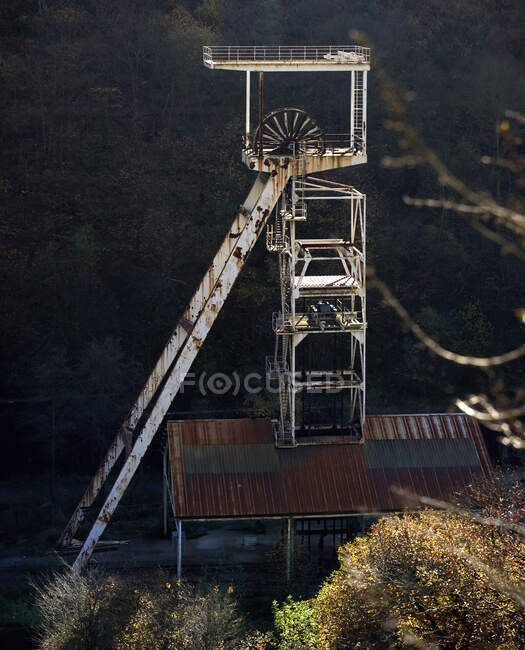 Deserted old tall rusty metal mine mechanism locating on background of blurred dark forest in countryside on sunny autumn day — Stock Photo