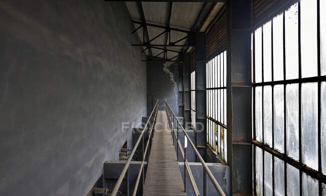 Schmale Holzbrücke mit Stahlgeländern über Räumen mit grauen Betonwänden und staubigen, schmutzigen, großen Fenstern in verlassenen Fabriken — Stockfoto