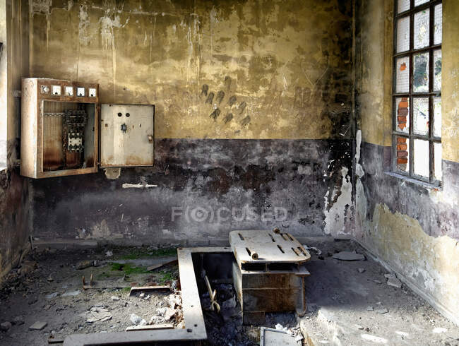 Разрушенная грязная комната со слоем пыли и кусочками бетона на полу, покрытом разбитым мохом, и старым неисправным трансформатором, висящим на потрепанной стене с черными пятнами в заброшенном здании — стоковое фото