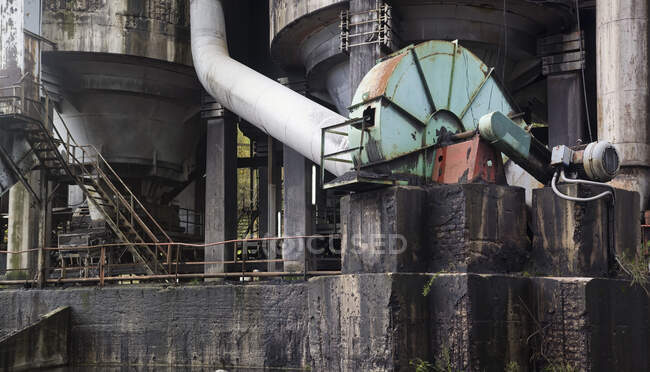 Часть огромного ржавого стального механизма, стоящего на грязных кирпичных черных блоках рядом с большими цилиндрическими структурами на заброшенном пустынном заводе — стоковое фото