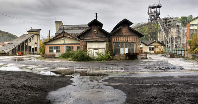 Мокрый неровный асфальт и потрепанный старый одноэтажный заброшенный деревянный дом на фоне зеленых деревьев и заброшенной угольной шахты в облачный прохладный день после дождя — стоковое фото