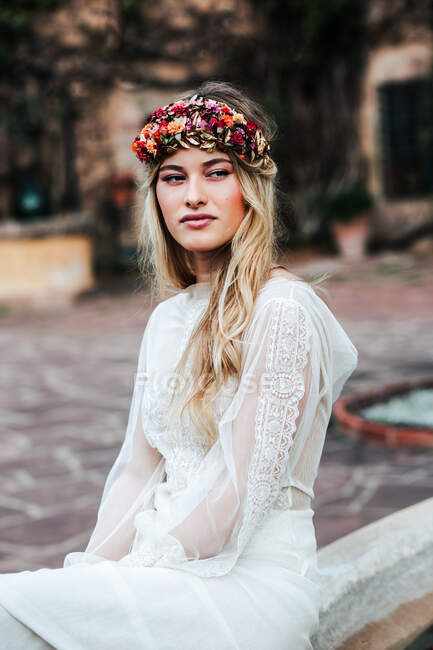 Hübsche junge Frau in transparentem weißen Kleid und Blumenkranz schaut weg, während sie am Hochzeitstag auf verschwommenem Hintergrund des Hofes sitzt — Stockfoto