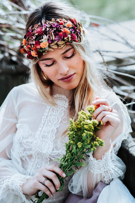 Belle jeune femme en robe blanche et couronne florale touchant bouquet de petites fleurs jaunes tout en se reposant dans le jardin le jour du mariage — Photo de stock