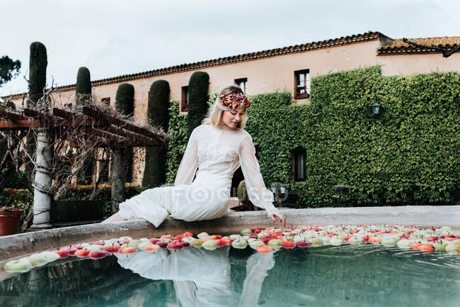Belle jeune femme en élégante robe blanche et couronne florale assise sur le bord sur le bassin de la fontaine de ciment et touchant pommes flottantes lors de la cérémonie de mariage dans le jardin — Photo de stock