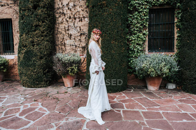 Side view of young woman in elegant white dress and floral wreath looking at camera while standing near aged house decorated with vines and potted bushes on wedding day — Stock Photo