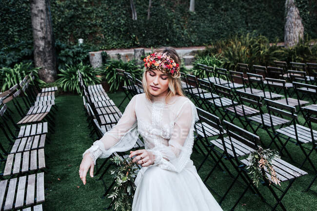 Hohe Winkel der schönen jungen Frau in elegantem weißen Kleid und Blumenkranz schließen die Augen und sitzen auf leeren Stühlen vor der Trauung im Garten — Stockfoto